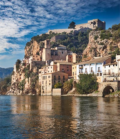 Attracties Foto genomen vanaf de rivier de Ebro met huizen langs de rivier en het kasteel Miravet op de top van de heuvel met uitzicht over de rivier.