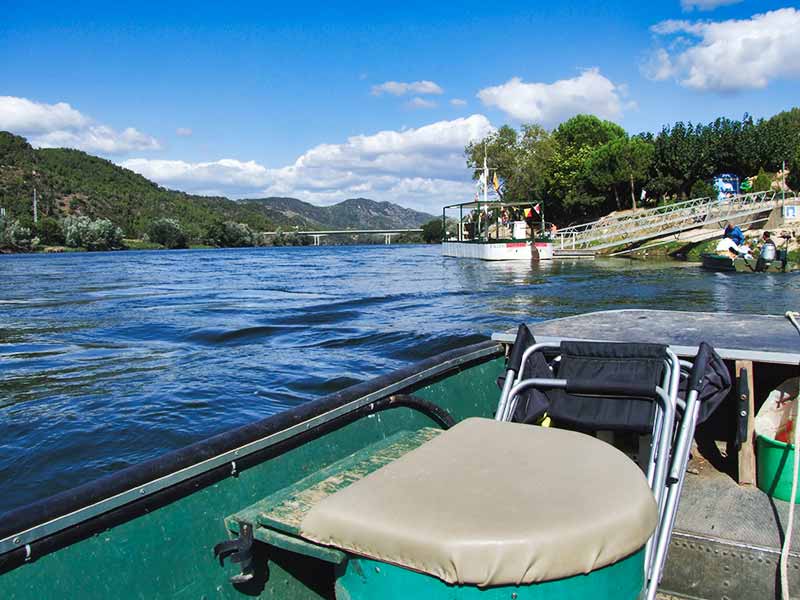 Vacaciones de pesca independientes o guiadas en el Ebro: ¿Cuál es el más adecuado para ti? Foto tomada desde el interior de un barco en el muelle de Benifallet, en el río Ebro, con vistas del puente de Benifallet y las montañas al fondo, bajo un cielo azul.