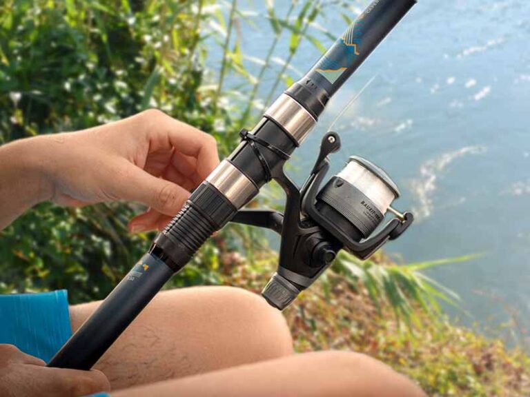 Pesca en el Ebro para Principiantes: Lo Que Necesitas Saber Foto de las manos y rodillas de un hombre que sostiene una caña de pescar y un carrete con el río Ebro en la Riverside Finca de Benifallet al fondo
